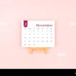 november 2027 calendar page with wooden easel on pink color background 3E4PP3M